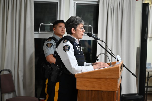 Insp. Kathy Hartwig stands indoors at a wooden podium speaking into a microphone with Staff Sgt. Major David Douangchanh standing beside her. 