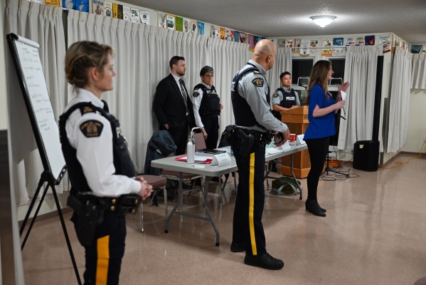 Four uniformed RCMP officers and Sgt. Tim Pierroti with IHIT stand near a table in a community hall looking on as Michelle Callander with Burnaby RCMP Victim Services speaks during a community outreach session