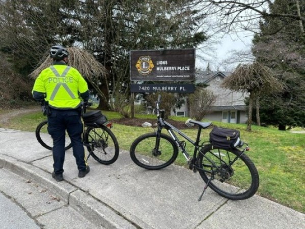 A Burnaby RCMP officer in a reflective yellow jacket and helmet stands outdoors beside two police bikes and a sign for Lions Mulberry Place