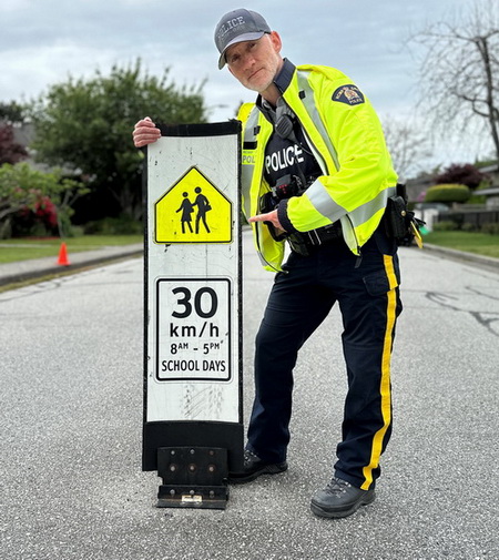 Photo d'un policier avec un panneau sur lequel il est écrit : « 30 km/h de 8 h à 17 h les jours d’école ». 