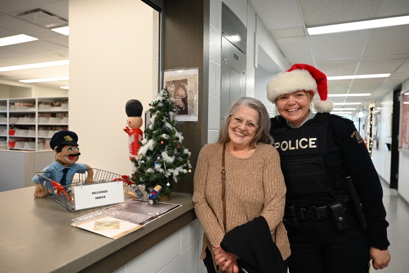 A police officer in uniform smiles beside a woman wearing a brown sweater indoors beside a small Christmas tree