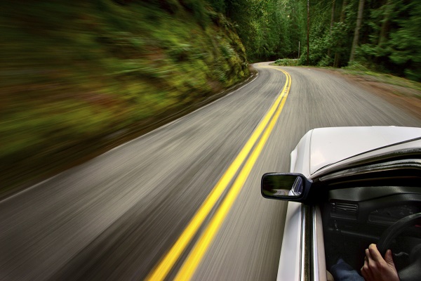 Exterior view of driver's door and mirror of vehicle speeding through a forest on a rural road.