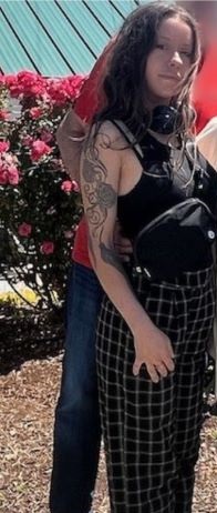 A woman wearing black and white checked pants, long brown hair, black tank top with a black belt bag, headphones around her neck and a large flower tattoo on her upper right arm