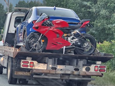 A red Ducati motorcycle is behind a blue sedan on the flat deck of a tow truck