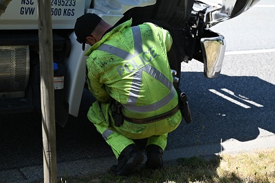 A police officer in a yellow reflective jumpsuit kneels down near the front tire of a commercial truck