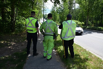 A bylaws officer stands beside two police officers in reflective yellow uniforms beside a roadway