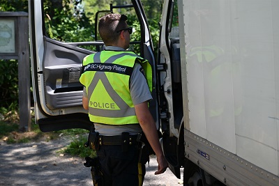 A police officer in reflective yellow uniform bends beside a commercial truck 