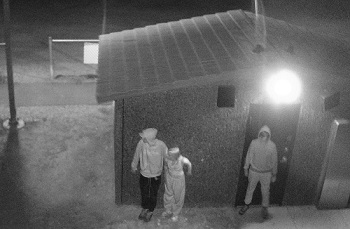 Photo en noir et blanc captée à l’aide d’une caméra de surveillance et montrant trois individus adossés au mur d’une structure abritant les toilettes publiques.