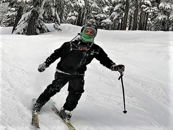 suspect male dressed in dark jacket and ski pants on the ski hill. He is wearing a helmet with a bright green neck/face warmer and holding ski poles.