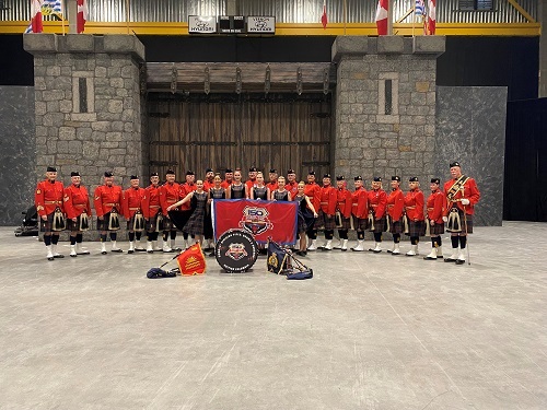 Photo de l’Orchestre de cornemuseurs, de tambours et de danseurs de la Division E derrière une bannière du 150e de la GRC. 