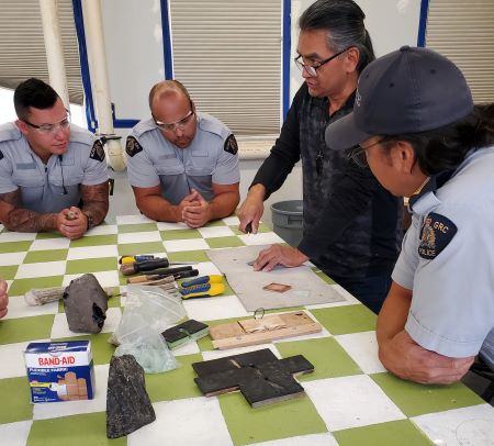 A group of uniformed police officers gathered around Ed Jensen while he demonstrated flint knapping on a checkered green and white table. Bandages, tools, and other supplies were on the table. 